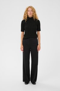 Žena s kučeravými blond vlasmi stojí oblečená v čiernom krátkom rukáve roláku, širokých čiernych nohaviciach a čiernych topánkach na bielom pozadí.