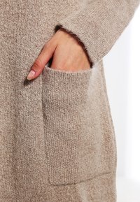 Swaeter w kolorze taupe z dzianiny, z widoczną boczną kieszenią i teksturowanym materiałem. Dół ma lekko luźny krój, a mankiety są prążkowane.