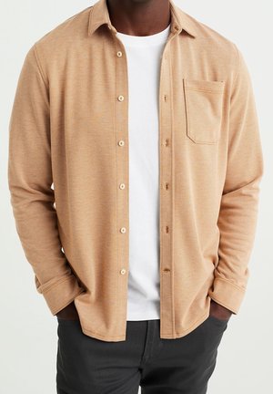 Mand iført en beige tekstureret skjorte med knapper over en hvid t-shirt med hænderne i lommerne og sorte bukser.