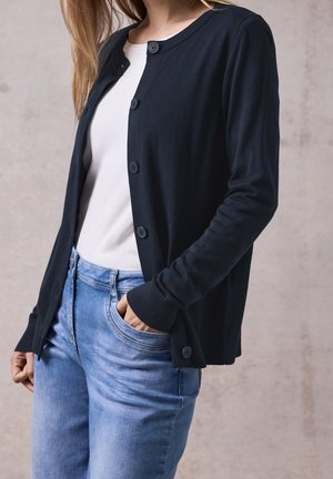Femme portant un cardigan bleu foncé boutonné sur un top blanc et un jean bleu clair, avec une main dans la poche du jean, sur un fond uni.