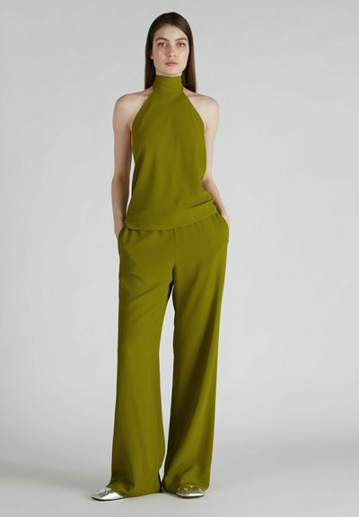 Haut en olive à col halter associé à un pantalon large. Le haut possède un décolleté haut, tandis que le pantalon a une coupe décontractée. Le mannequin porte des chaussures argentées.