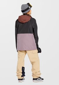 Schneejacke in Schwarz, Braun und Lavendel, mit Kapuze, kombiniert mit beigen Cargo-Hosen mit schwarzen Akzenten und schwarz-weißen Sneakers.
