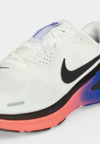 Λευκό αθλητικό παπούτσι Nike με μαύρο σήμα (swoosh), κοραλλί και μπλε σόλα, πλεκτό επάνω μέρος και ετικέτα «Structure 26» στη γλώσσα.