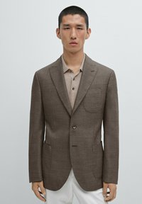 Brązowa wciąż blazer z wyciętym kołnierzem, pojedynczym guzikiem, dwoma przednimi kieszeniami i dopasowanym krojem, w połączeniu z jasnobrązową koszulą z kołnierzykiem.