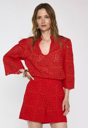 Femme aux longs cheveux portant un chemisier rouge à motif œillet et une jupe assortie, posant avec une main sur la hanche sur fond blanc.