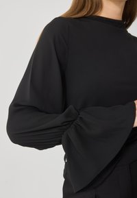 Blusa nera con maniche lunghe e svasate, caratterizzata da dettagli arruffati ai polsi. Tessuto liscio con una lieve lucentezza. Collo alto, senza motivi visibili.