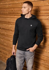 Black hoodie with a front pocket and a small logo patch, paired with light gray jeans, against a wooden backdrop.