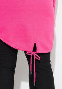 Tunique rose avec un ourlet arrondi et un cordon coulissant sur le côté, assortie à un pantalon noir. Le cordon dispose de terminaux en argent.
