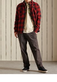 Camisa de franela de cuadros rojos y negros sobre una camiseta beige, combinada con pantalones grises y zapatillas negras. Fondo de paneles de madera.