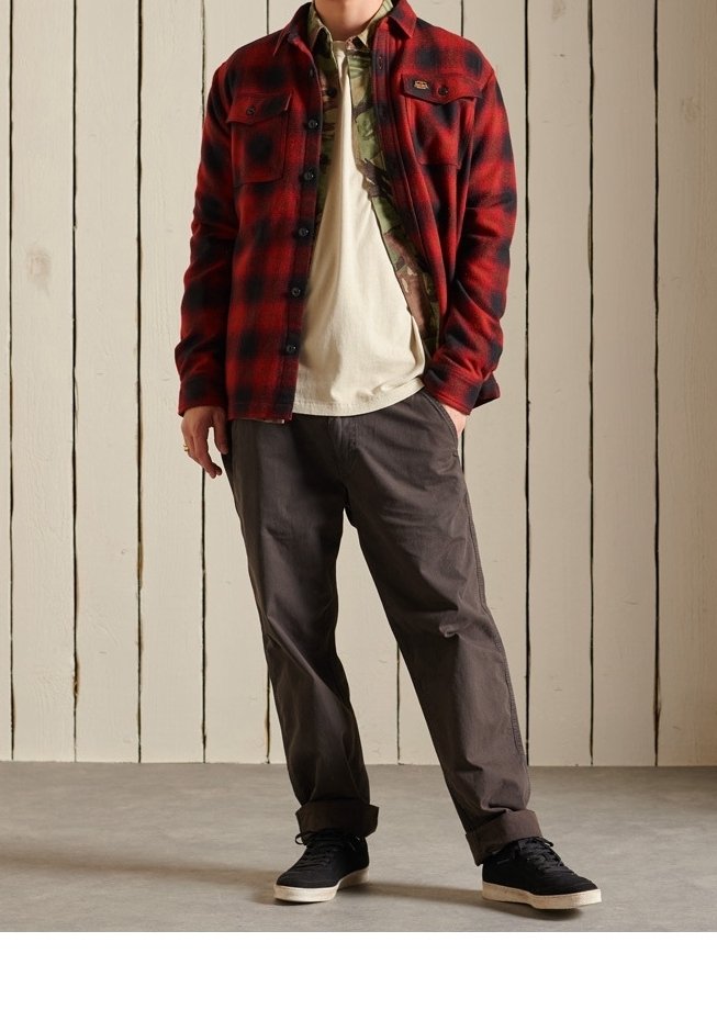 Camisa de franela de cuadros rojos y negros sobre una camiseta beige, combinada con pantalones grises y zapatillas negras. Fondo de paneles de madera.