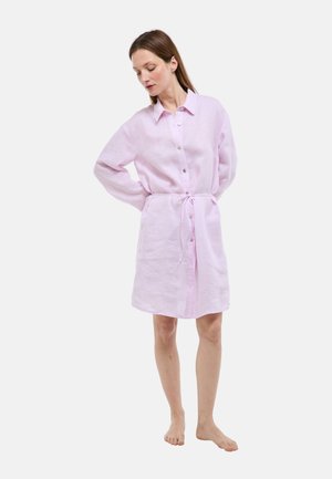 Femme debout pieds nus, portant une robe boutonnée à manches longues rose clair avec une ceinture à la taille, regardant vers le bas à sa gauche.
