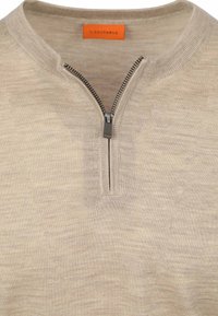 Beige ritsvest gemaakt van zachte stof. Heeft een opstaande kraag en een zilveren rits aan de voorkant. Het label heeft een oranje achtergrond met zwarte tekst.