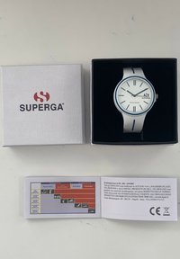 Reloj de silicona blanco con acentos azules, cara redonda y marcadores de hora plateados. Incluye una caja blanca etiquetada como "SUPERGA" y un folleto informativo.