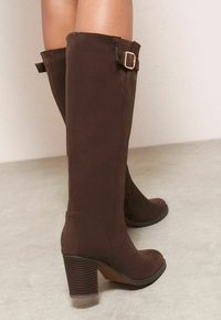 Botas altas hasta la rodilla de ante marrón con tacón de bloque, que presentan una hebilla decorativa y detalles de costura a lo largo del lateral.