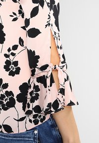 Blusa rosa chiaro con motivo floreale nero, dotata di maniche a volant e dettagli a intaglio. Realizzata in tessuto morbido con una vestibilità rilassata.