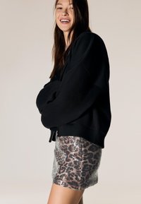 Črni oversized sweatshirt z širokimi rokavi, v kombinaciji z bleščnim mini krilom z leopardskim vzorcem, narejenim iz sequins. Nevtralen ozadje.