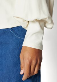 Cream-colored, soft fabric top with voluminous sleeves. A hand rests near a blue denim garment, showcasing smooth texture and white stitching.