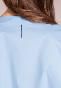 Indumento azzurro chiaro in tessuto morbido, con una striscia verticale di decorazioni scure sul colletto posteriore.