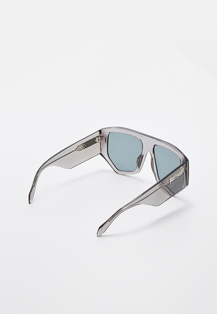 Lunettes de soleil avec une monture grise transparente, des verres carrés angulaires et une subtile teinte bleue. Branches intérieures texturées avec un détail de marquage.
