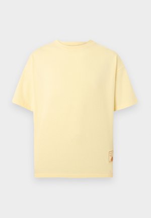 T-shirt à manches courtes de couleur jaune clair avec un col rond, doté d'une petite écusson rectangulaire près de l'ourlet inférieur sur le côté gauche.