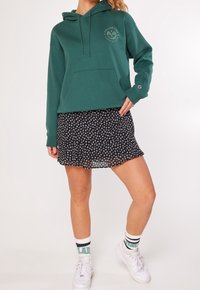 Kvinna i en grön Brooklyn Tennis Club hoodie, svart blommig minikjol, vita sneakers och vita strumpor med gröna och svarta ränder.