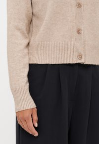 Bež kardigan z rebrastimi manžetami in robom, ki ima šest svetlo rjavih gumbov. Mehka tekstura dopolnjuje črne hlače, ki so vidne spodaj.