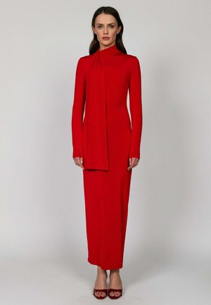 Kobieta stojąca w długiej czerwonej sukience z wysokim dekoltem i długimi rękawami oraz dopasowanych czerwonych butach na obcasie z odkrytymi palcami na jednolitym tle.