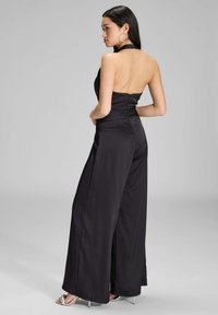 Schwarzer Satin-Overall mit weitem Bein, Neckholder und gerüschtem Detail auf der Rückseite. Verfügt über einen verdeckten Reißverschluss und eine glatte Textur.