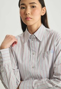 Chemise à manches longues à rayures avec des lignes bleues, rouges et blanches, dotée d'une poche poitrine et d'une texture douce. Arrière-plan gris clair.