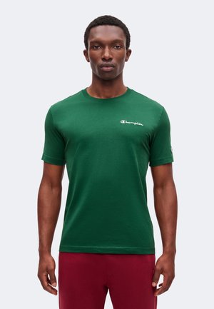 Hombre vestido con una camiseta Champion verde de manga corta y pantalones rojos, de pie frente a un fondo claro y sencillo.