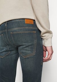 Mörkblå denimjeans med en slät textur, med en brun läderlappar bak och orange dekorativ söm på fickorna.