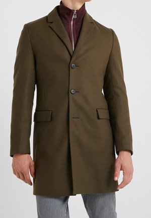 Homme portant un manteau vert olive cintré avec deux boutons noirs et des poches à rabat, sur un haut zippé bordeaux et un pantalon rayé.