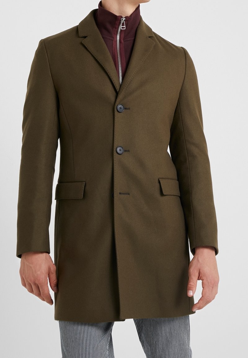 Homme portant un manteau vert olive cintré avec deux boutons noirs et des poches à rabat, sur un haut zippé bordeaux et un pantalon rayé.