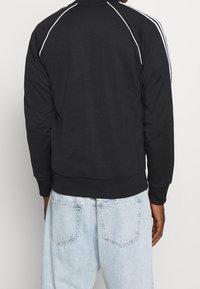 Chaqueta negra con cierre y costuras blancas, puños y dobladillo acanalados, hecha de un tejido suave. Combinada con unos vaqueros holgados de color azul claro.