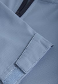 Ljust blått tyg med en slät yta har en kardborrestängning och en svart dragkedja. Designen inkluderar subtila rutmönster och noggrant sydda sömmar.