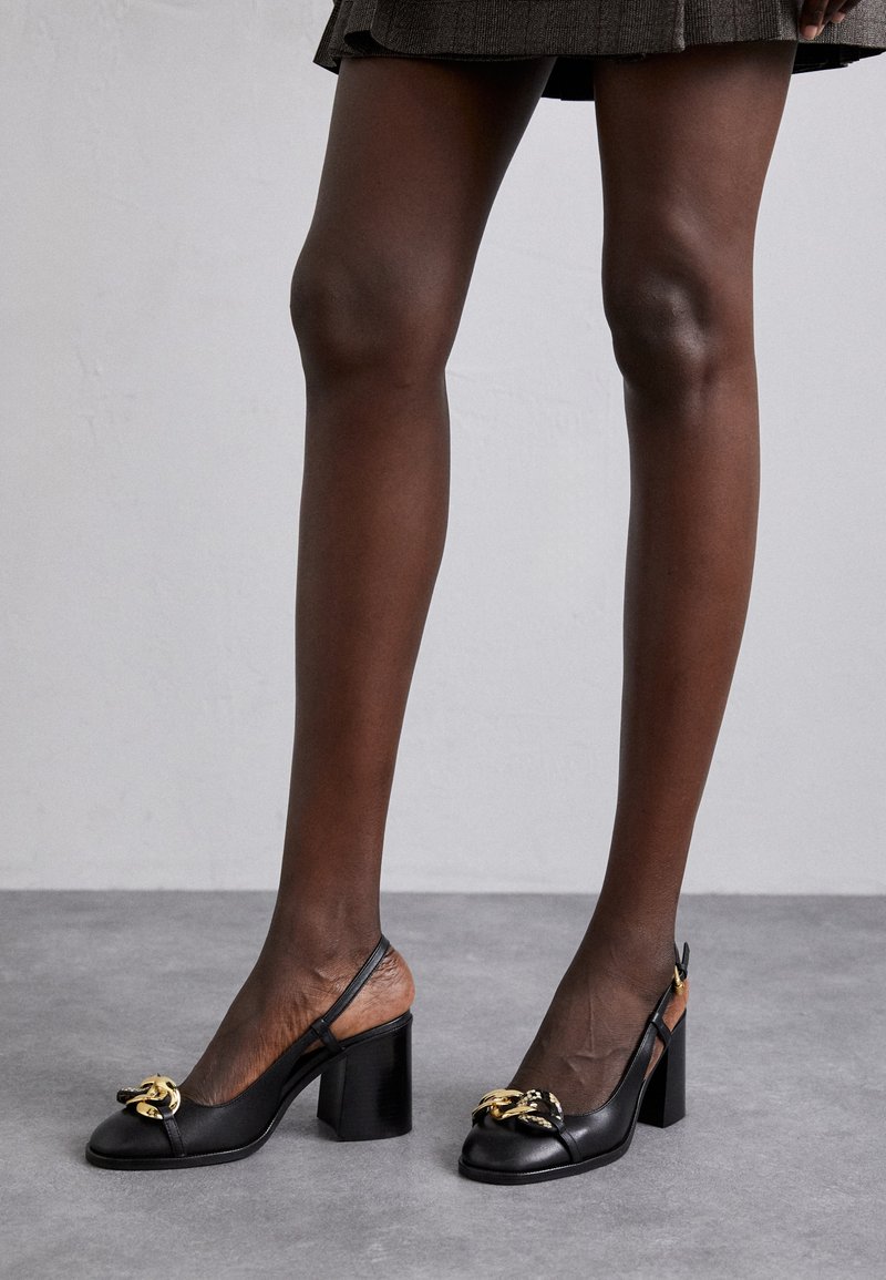 Legs wearing a short plaid skirt and black slingback block heels with gold chain detail, standing on gray floor against light wall.