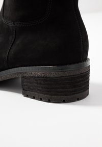 Primer plano de una bota de tobillo de ante negro, con un tacón de bloque texturizado y una superficie estriada, junto con acentos negros suaves a lo largo de la suela.