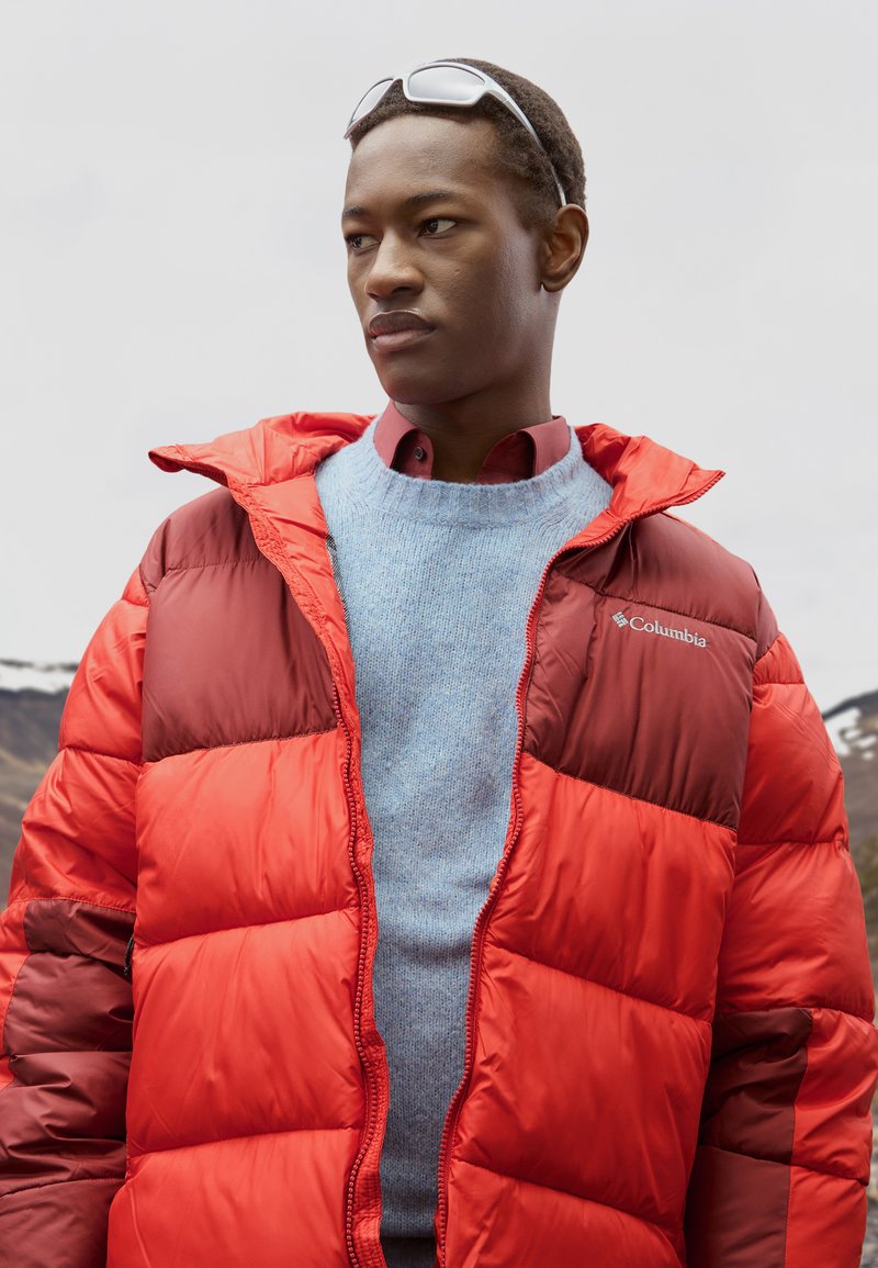 Joven con chaqueta acolchada roja de Columbia y suéter azul, mirando a la izquierda con gafas de sol en la cabeza, paisaje montañoso al fondo.