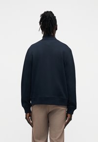 Μπλε ναυτικό φούτερ με χαλαρή εφαρμογή, ριμπ μανσέτες και τελείωμα, φορεμένο πάνω από ανοιχτό καφέ παντελόνι με ραφή, φωτογραφημένο από πίσω.