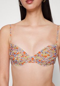 Sutiã com estampa floral e aros, apresentando alças finas e ajustáveis. As cores predominantes incluem roxo, laranja e amarelo sobre um fundo claro.