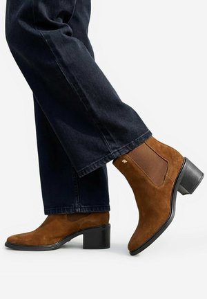 Braune Wildleder-Stiefeletten mit Blockabsatz, getragen zu dunkelblauen Jeans vor weißem Hintergrund, mit Seiten- und Rückansicht.