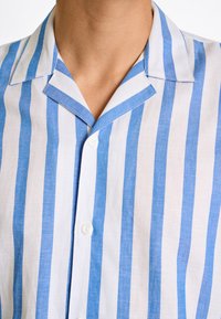 Camicia a righe blu e bianche realizzata in tessuto leggero, con colletto a punte e bottone frontale. Strisce orizzontali distintive.