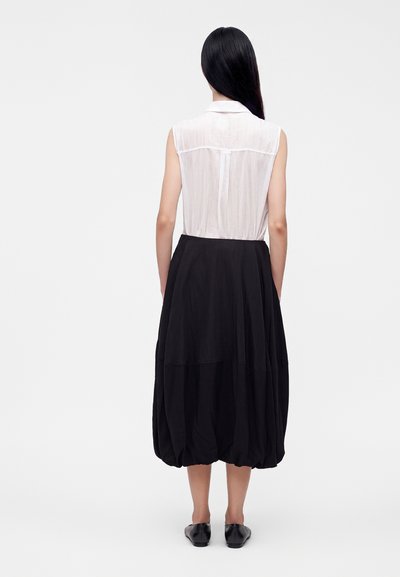 Femme aux longs cheveux noirs portant un chemisier blanc sans manches et une jupe midi noire ample, debout, dos tourné, sur un fond uni.