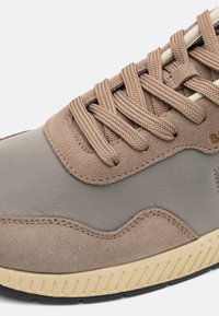 Dessus en suède marron et synthétique gris, avec des lacets plats et une semelle extérieure en caoutchouc beige. Présente une couture ondulée et des détails de couture.