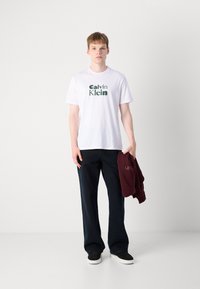Hvit bomulls t-skjorte med grønn "Calvin Klein" logo, mørkeblå vide bukser og en bordeaux hettegenser drapert over den ene armen. Svarte sneakers.