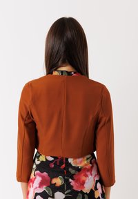 Giacca corta marrone con maniche a tre quarti, design strutturato, texture liscia, indossata sopra un vestito a fantasia floreale dai colori vivaci.