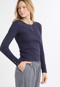 Marineblaues Henley-Oberteil mit langen Ärmeln und einer gerippten Textur, mit rundem Ausschnitt und drei metallischen Knöpfen am Kragen, kombiniert mit gestreiften Hosen.