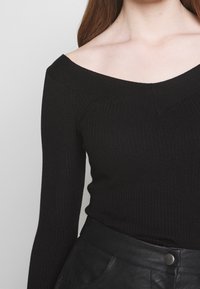 Zwart geribd gebreid topje met een off-shoulder ontwerp en lange mouwen, met een V-halslijn en een gladde textuur. Gecombineerd met zwarte leren broek.