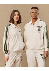 Two models wear coordinated cream athletic jackets with green stripes. The jackets feature a zip closure and "YALE" logo in black.