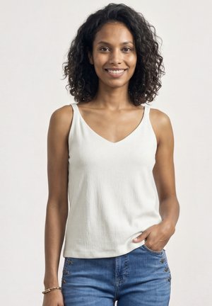 Smilende kvinde med krøllet hår iført en hvid ærmeløs top og blå jeans, stående med den ene hånd i lommen.